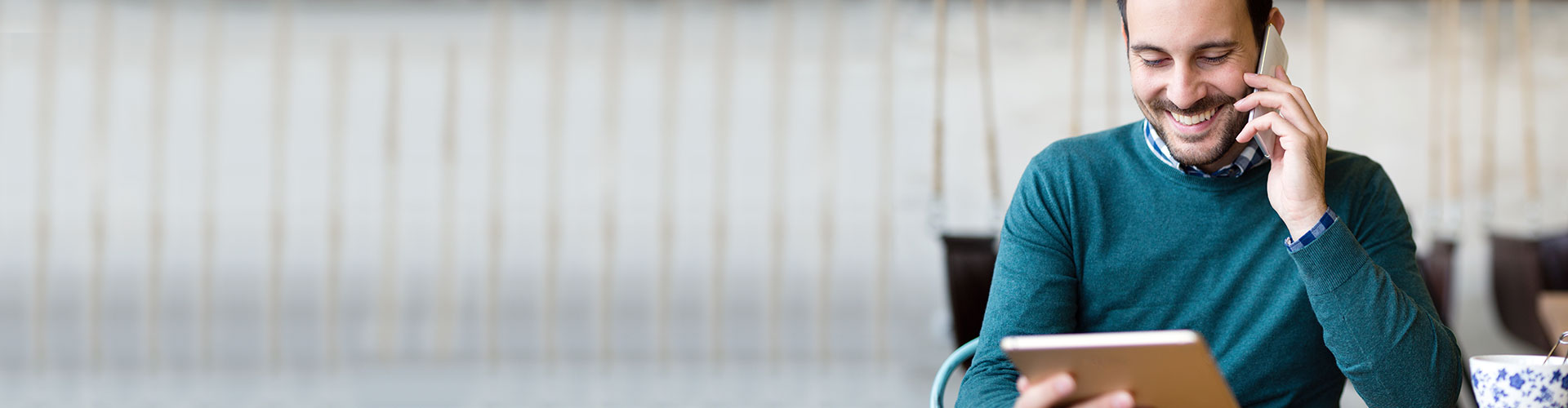 Empresas - Hombre de negocios con jersey azul sentado en una cafetería hablando por teléfono mientras navega por Ruralvía mi negocio en su tablet Empresas - Hombre de negocios con jersey azul sentado en una cafetería hablando por teléfono mientras navega por Ruralvía mi negocio en su tablet